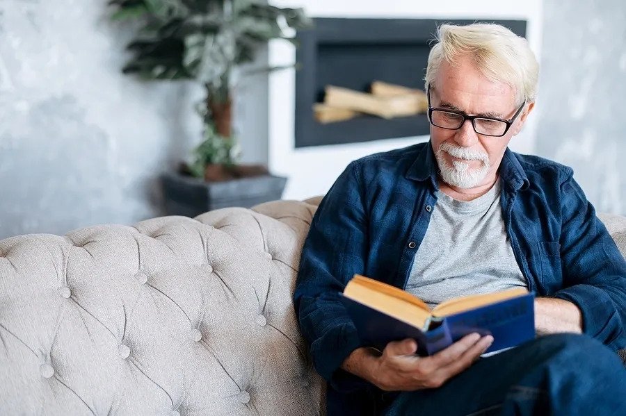 Senior man spends leisure time reading book sitting on the comfortable couch at cozy home.